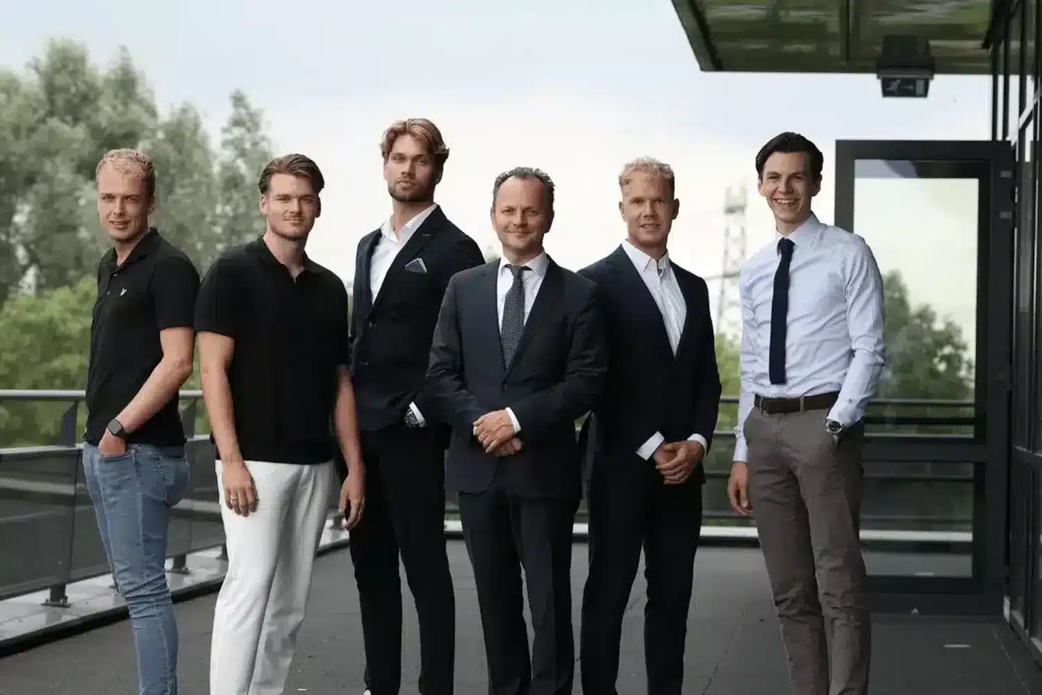 Groepsfoto van zes mannen die naast elkaar staan te glimlachen op een dakterras met een glazen kantoorgebouw op de achtergrond