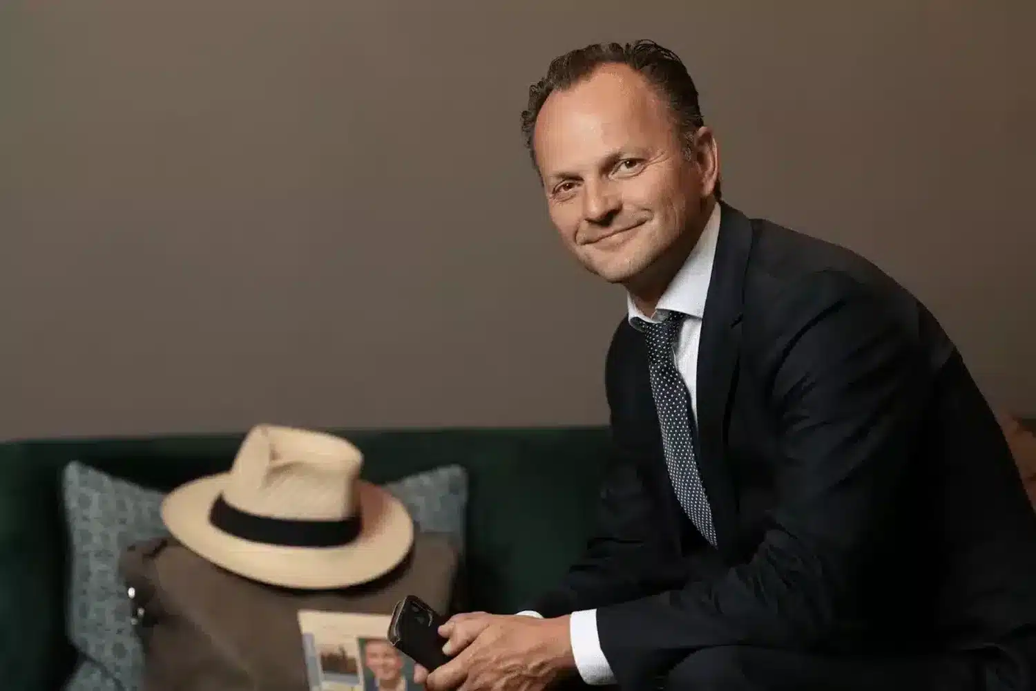 Foto van een Jan-Pieter in pak op een groene fluwelen bank met een smartphone en een panama-hoed naast hem