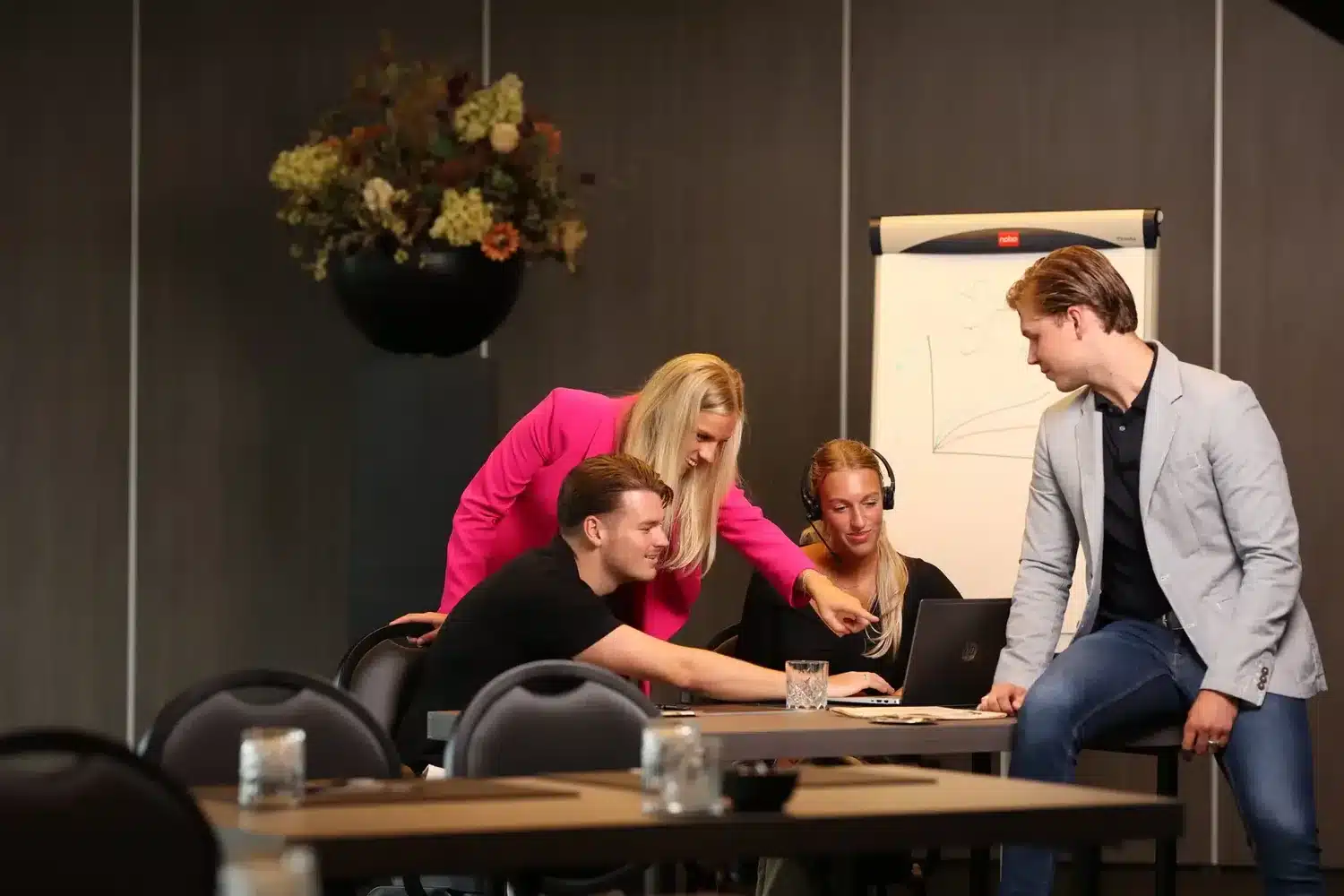 Foto van vier collega's rond een vergadertafel met laptop, vrouw in felroze blazer wijst naar het scherm
